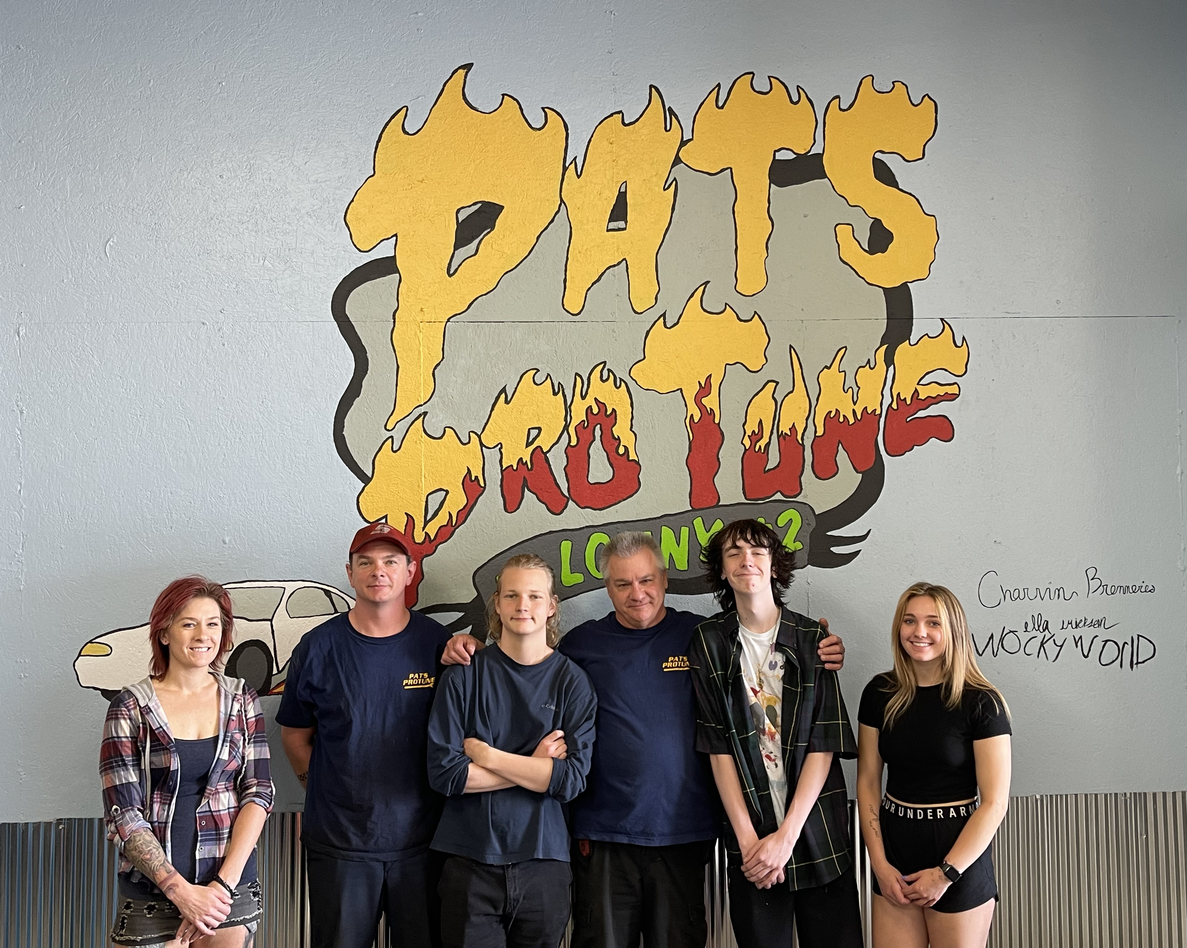 A group of four high school students pose in front of an auto repair shop mural with the business' two owners.