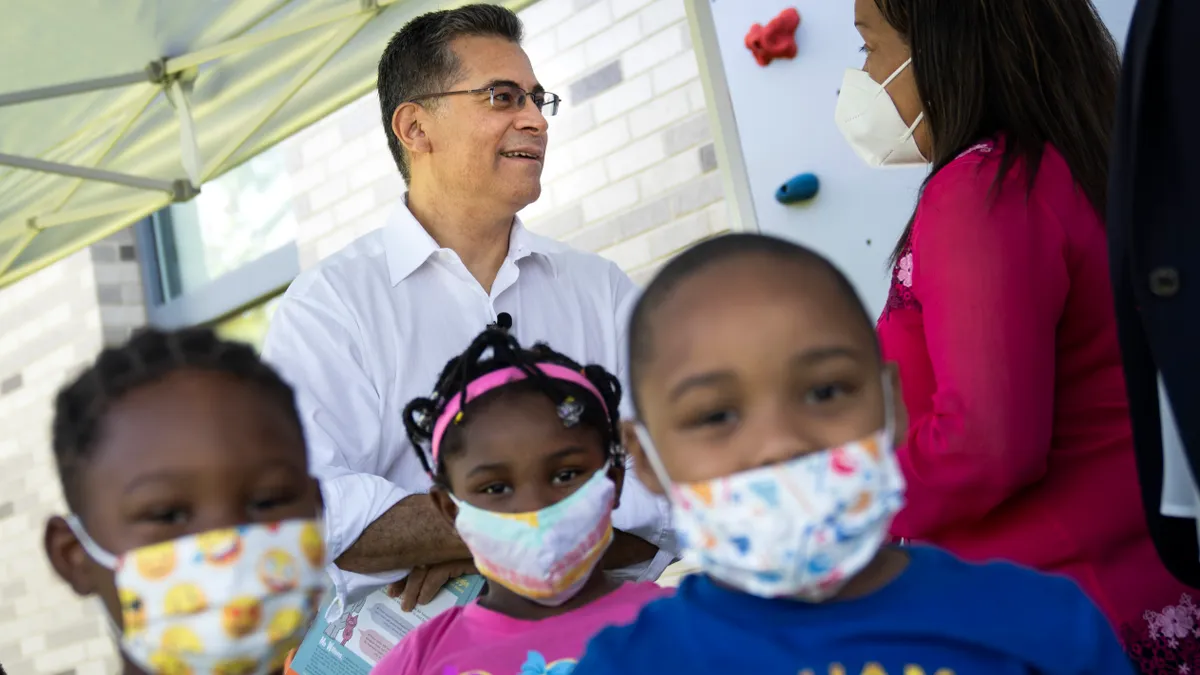 Two adults are pictured in the background looking at each other. Three young children are in the foreground wearing face masks and looking at the camera.