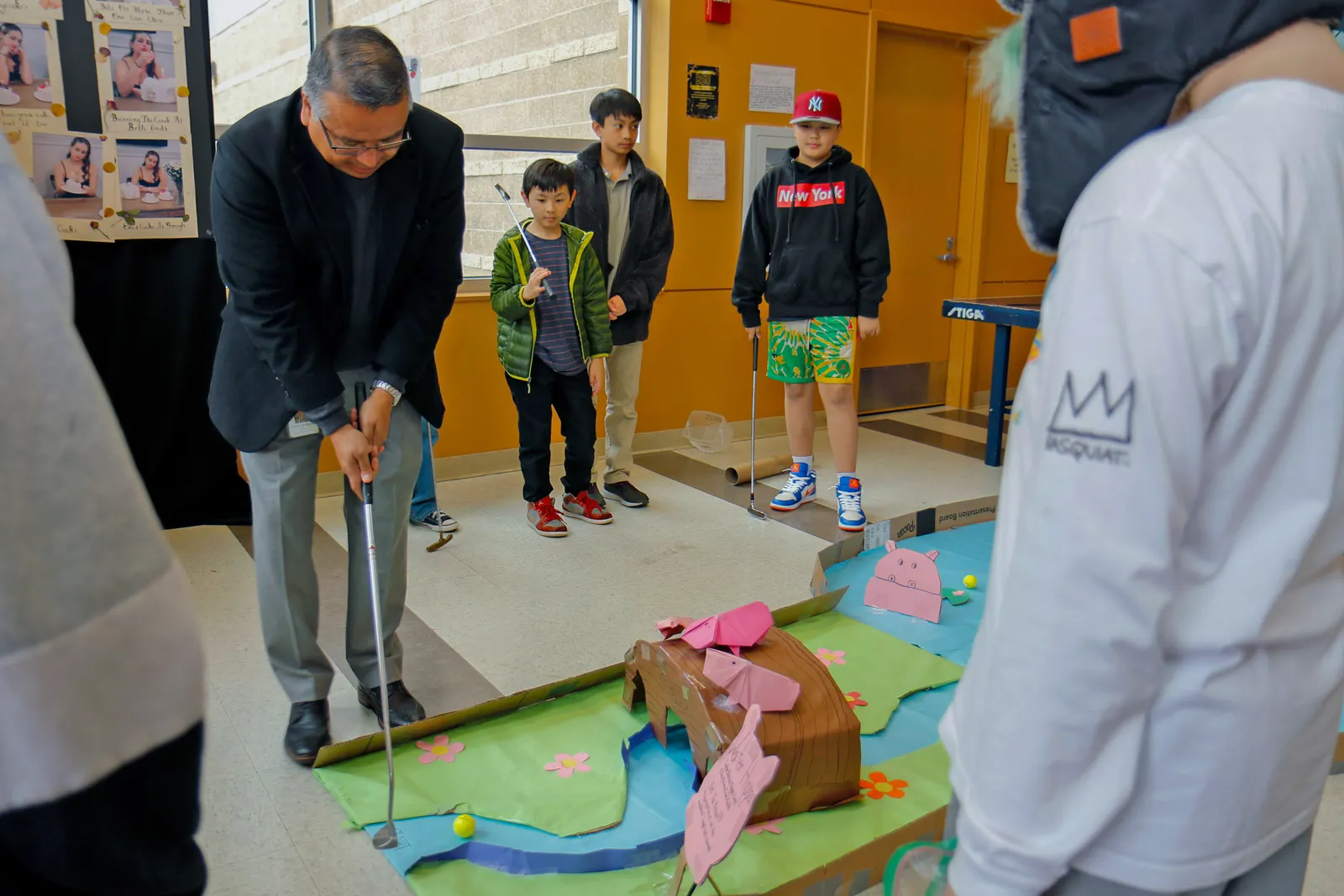 A person stands over a homemade mini golf hole holding a putter. Several students are standing and watching.