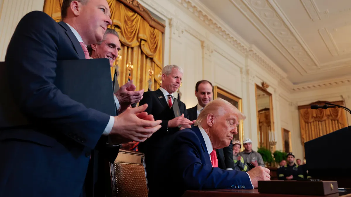 President Donald Trump sits at a desk signing a document with several people wearing suits standing behind him.