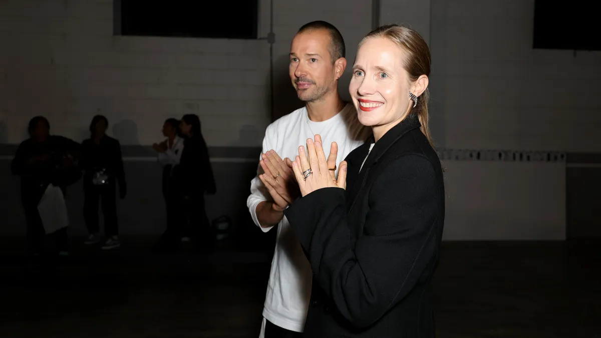 A man stands slightly behind a woman. Both clasp their hands in gratitude and angle themselves toward the camera.