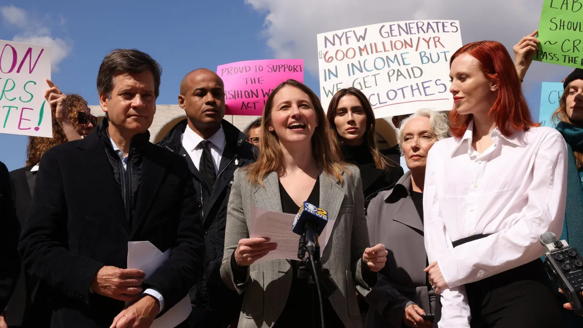 A group of people stand outdoors in support of the Fashion Workers Act. Several people hold signs, some reading "NYFW generates $600 million a year in income but we get paid in clothes." "Proud to support the Fashion workers act."