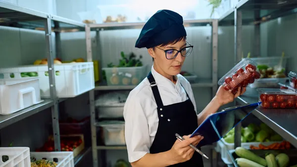 A person wearing a chef outfit holds up a container of tomatoes and looks at clipboard. The person appears to be in a kitchen storage area.
