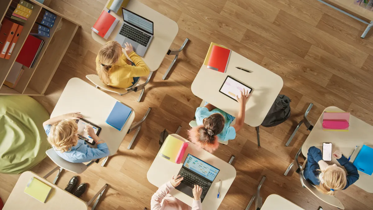 An above camera shot of students seated at individual desks in a classroom. On their desks are laptops or tablets.