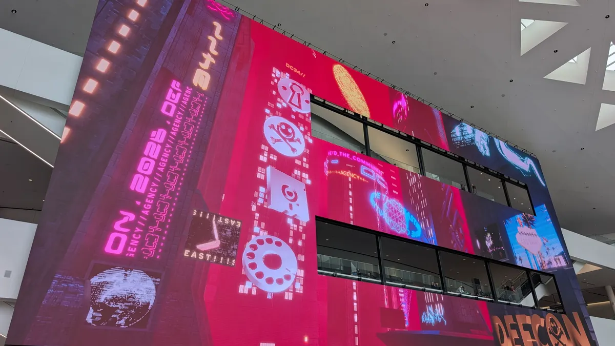 A large screen inside a convention center displays logos and other imagery related to the DEF CON cybersecurity conference