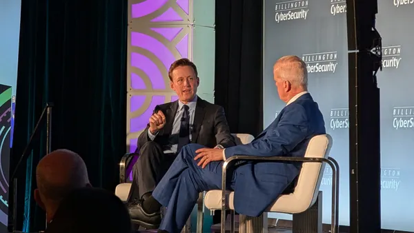 Two men wearing suits sit in white chairs on a stage