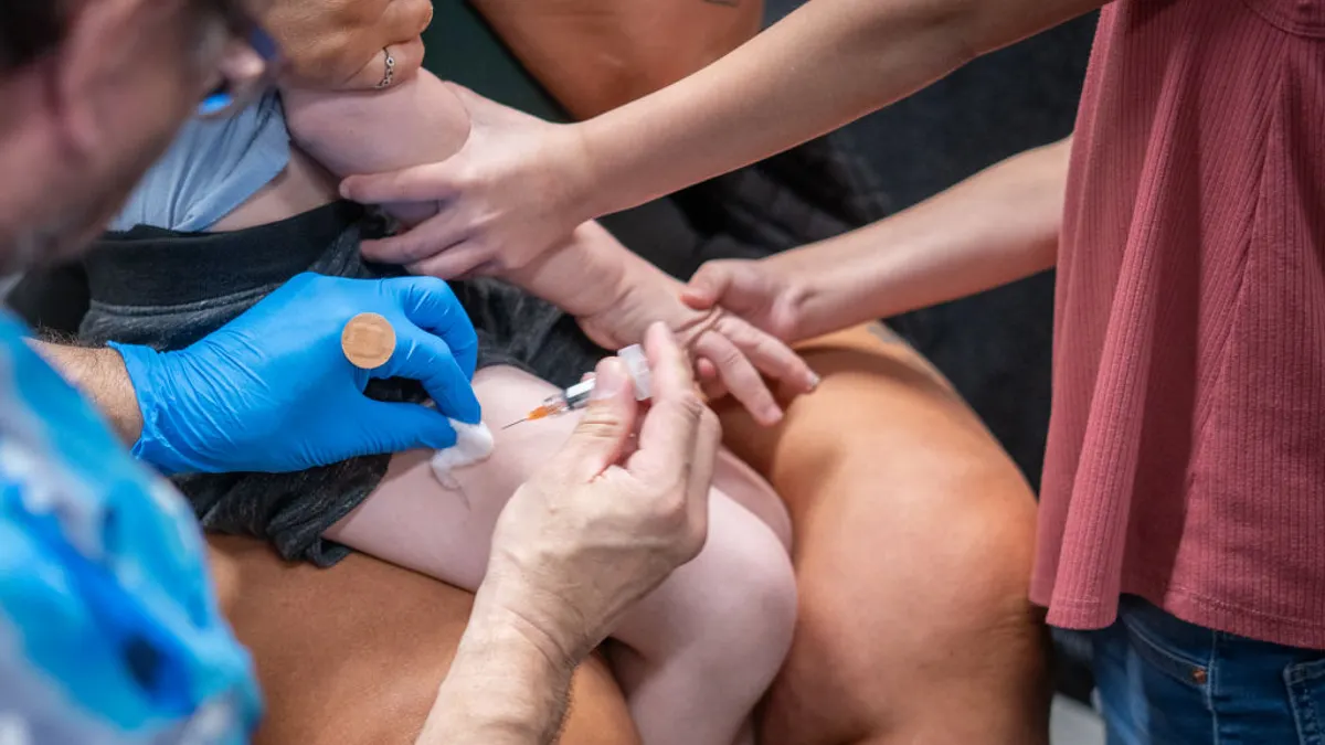 A syringe is being inserted into the leg of a child. A gloved hand holds a Baid-Aid. Another pair of hands seems to hold the arms of the child.