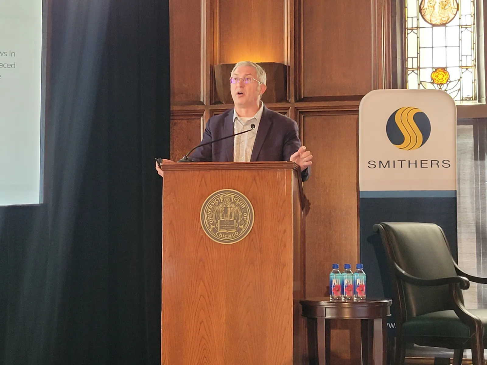 A person speaks while standing behind a podium behind a sign with the Smithers logo.