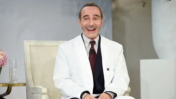 A man in a white suit smiles while sitting in a chair on stage.