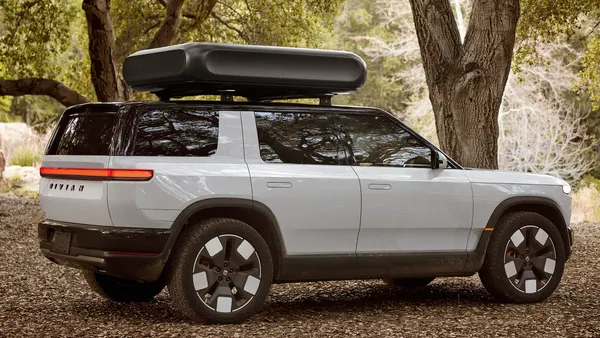 A white Rivian R2 electric SUV with a black cargo carrier on the roof parked outdoors with trees in the background.