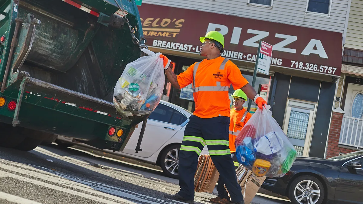 Two workers in orange shirts and yellow hard hats collect recyclables to throw into a truck, in front of a pizza restaurant in New York City