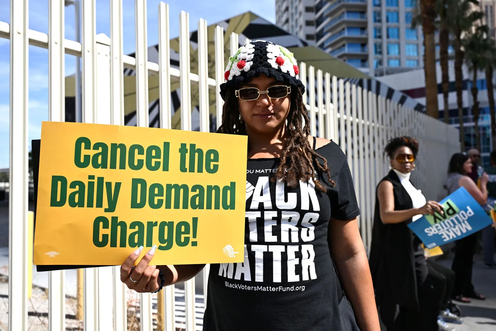 A person wearing black holds a sign reading "Cancel the daily demand charge!"