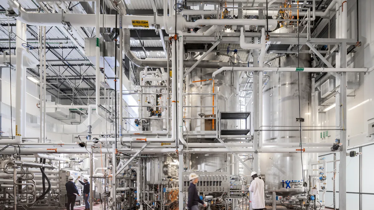 Machinery, including large tanks and piping, used for biomass fermentation in an industrial building with red tile floors. A few people in hardhats are near the equipment.