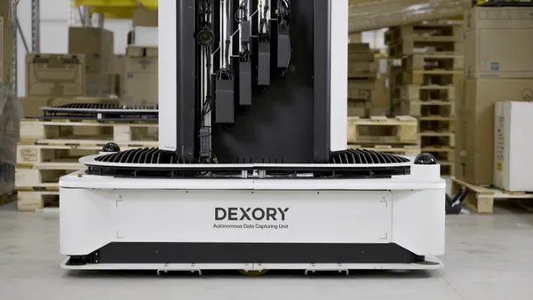 A white autonomous data capturing unit labeled “DEXORY” sits on a warehouse floor, with a tall black scanning structure mounted on top. Shelves, pallets and storage boxes are visible in the background.