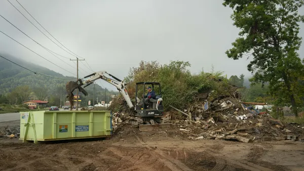 A man drives a small construction claw truck on a dirt lot near a road, transferring a large mound of wood waste and debris into a dumpster.