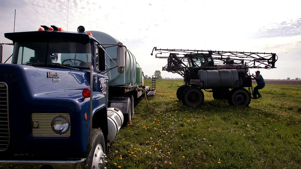 A farmer stands next to an herbicide sprayer. Next to him is a truck.