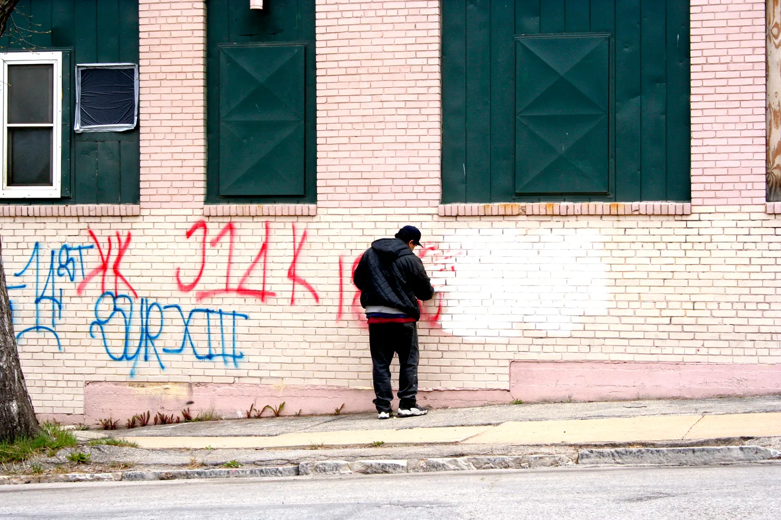 A person wearing a hooded jacket and sneakers applies white paint to the side of a building spraypainted with red and blue letters