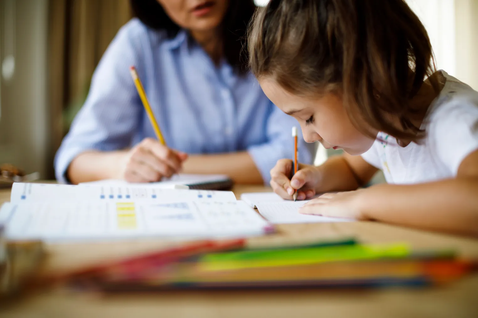 Student in foreground sits at a table with an adult. Both are holding pencils and there are workbooks on the table.