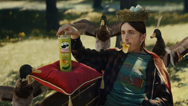 A person splays on a park bench with Pringles in his mouth as ducks flock around him
