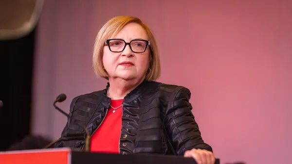 A woman, dressed in black and red, looks to the left as she stands at a black-and-red podium while speaking.
