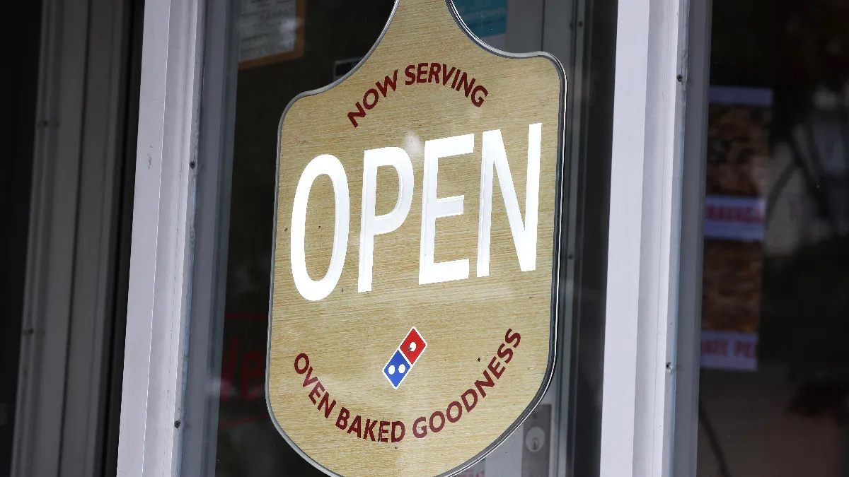 An image of a sign hanging on a window that says "Now Serving Open Oven Baked Goodness"