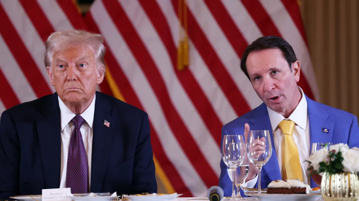 President Trump and Louisiana Gov. Jeff Landry sit at a table with a white tablecloth and two empty wine glasses with American flag behind them.