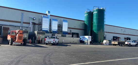 A flat industrial building across a paved roadway with a pair of tall dark green silos in the foreground and large silver vent tubes on the building.