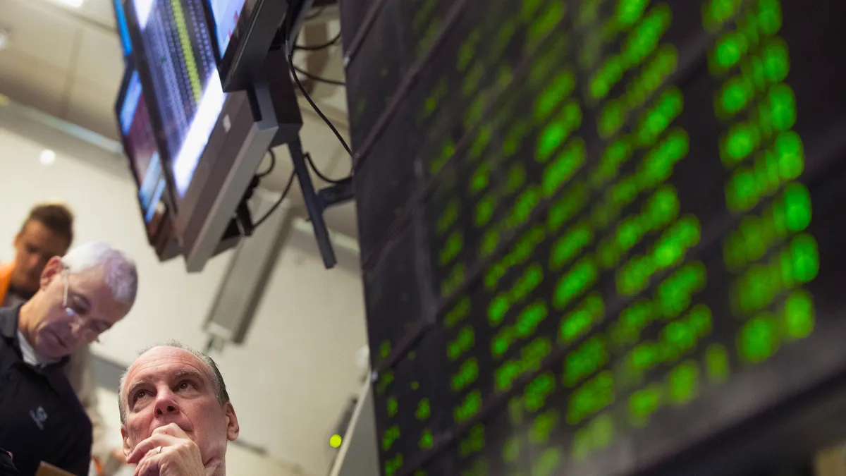 A stock trader with hand on chin in the bottom left looks up at market screens with out of focus TVs showing the S&P 500.