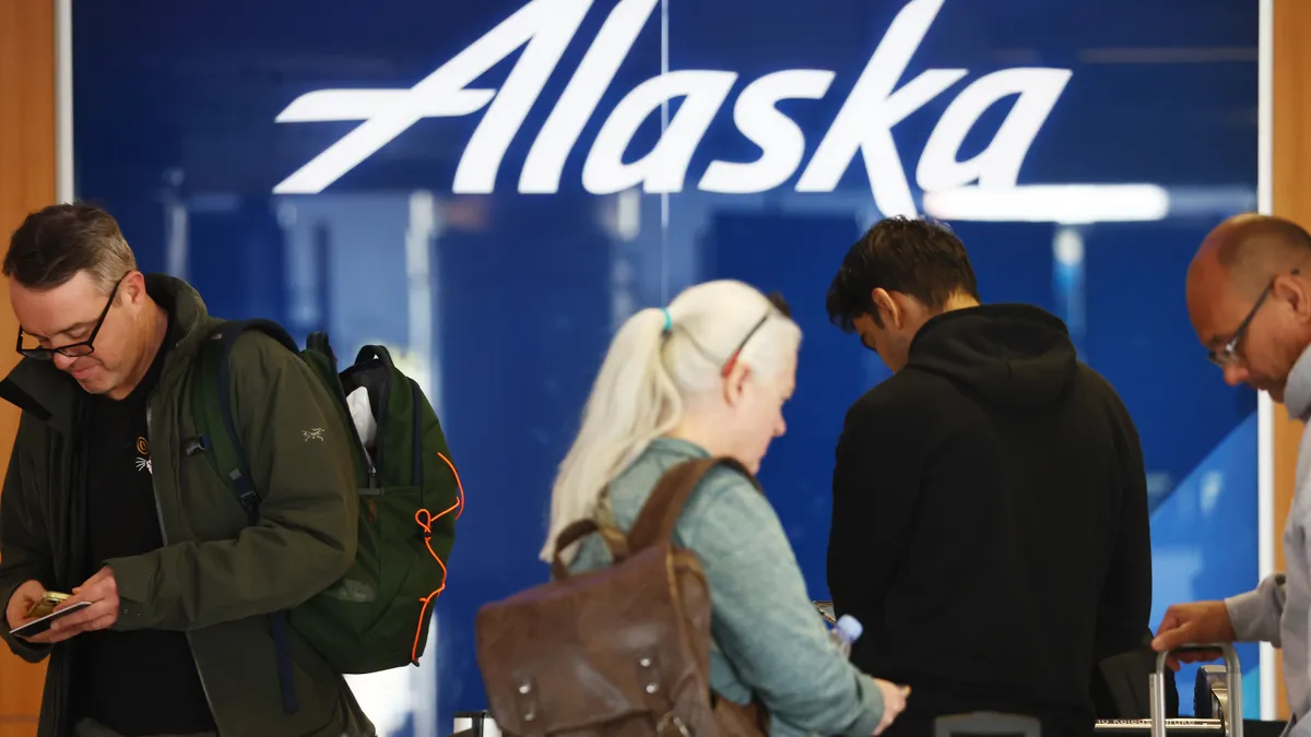 Travelers stand at a check-in area in front of an Alaska Airlines sign.