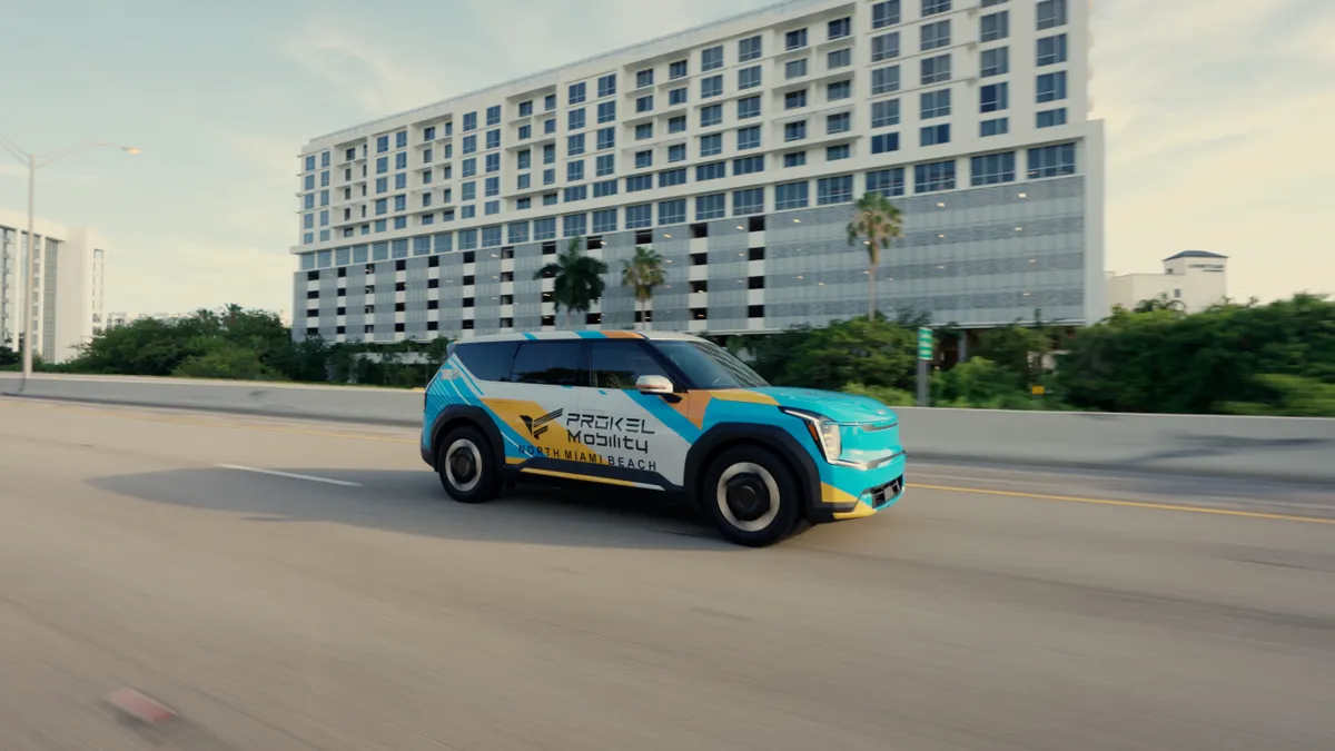 A green, blue and white SUV lettered "Prokel Mobility" on a road in front of a multistory building.