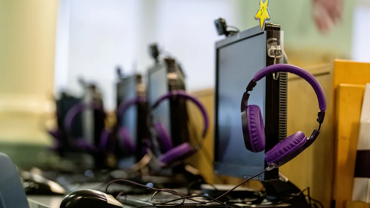 A school computer lab setup featuring purple headphones hanging on monitor stands with multiple desktop computers arranged in rows.