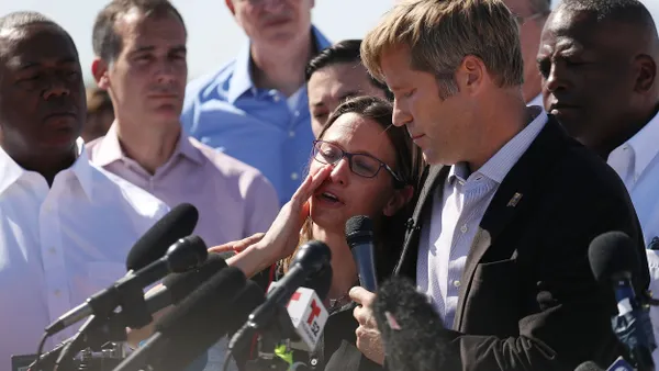 A person whispers into the ear of another person wearing a suit and speaking into several microphones, surrounded by many people.