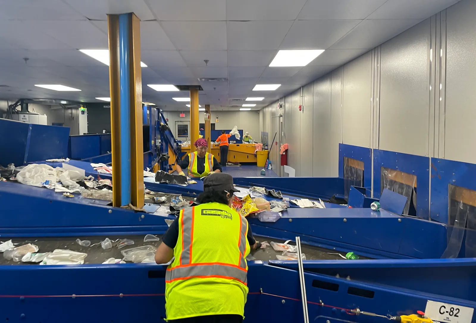 Workers in yellow vests stand over a conveyor belt sorting plastic recyclables