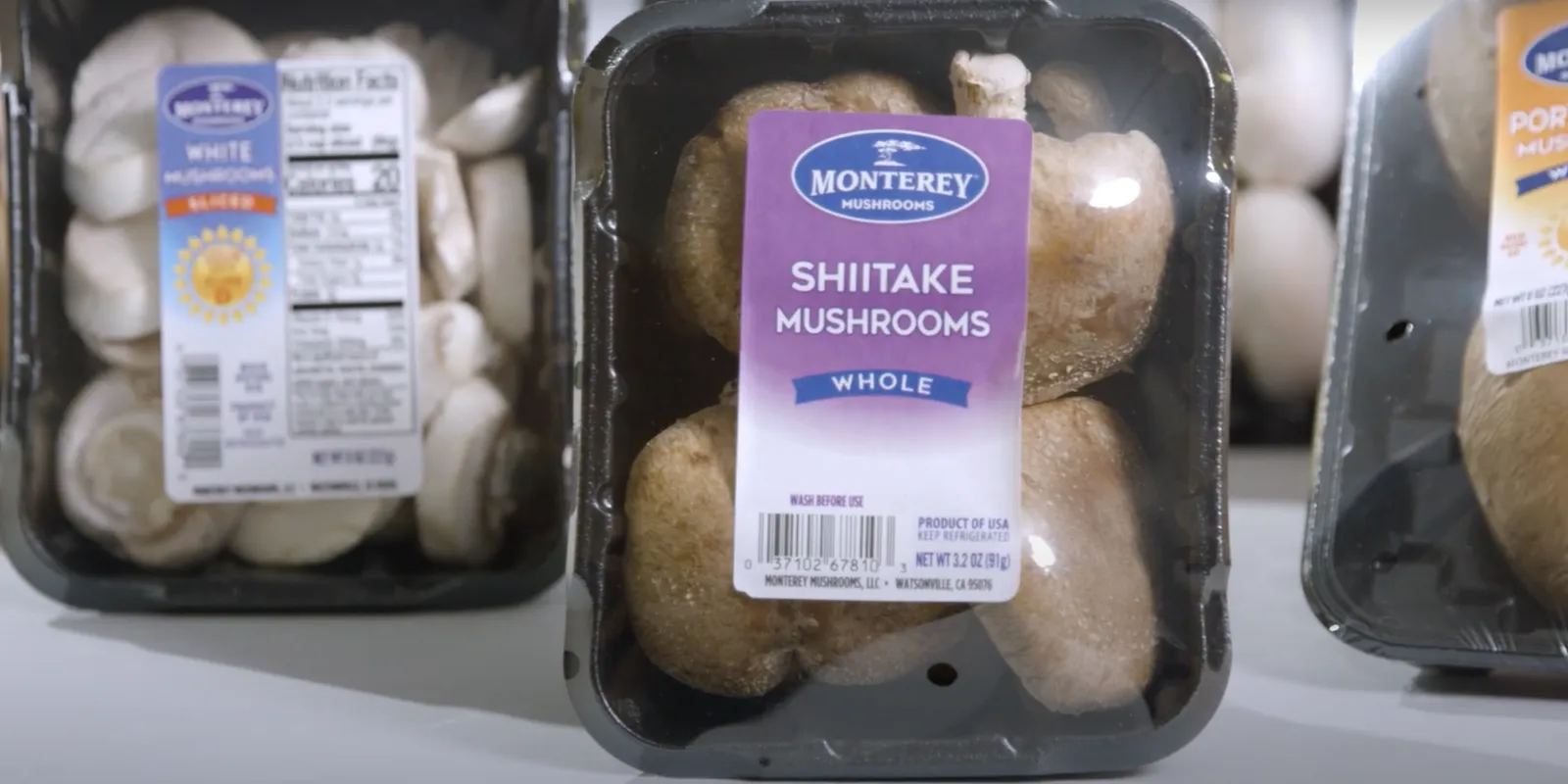 Three packages of different kinds of Monterey Mushrooms in black containers with clear plastic over the top.