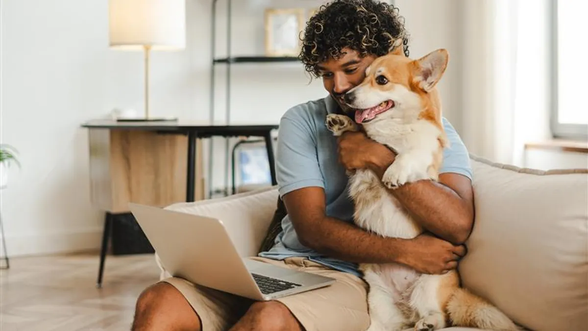 Man with curly hair sitting on a couch, hugging a smiling corgi. An open laptop rests on his lap. The room is warm and cozy, conveying a relaxed vibe.
