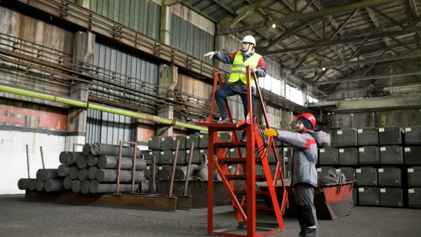 Two workers in safety gear operate in an industrial warehouse. One stands atop a red mobile ladder giving directions, while the other stabilizes the ladder from below. Stacks of cylindrical graphite rods and large blocks are visible in the background. The facility has a high ceiling with exposed beams and industrial lighting.