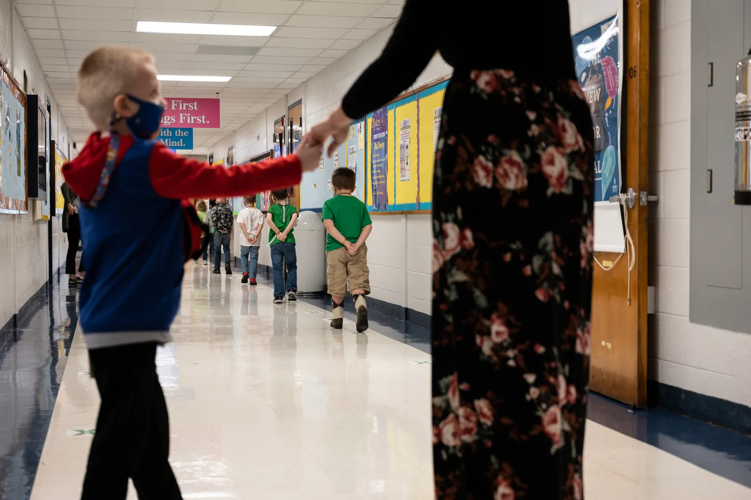 A young student wearing a face mask holds the hand of an adult in a school hallway as after young students walk down the hall.