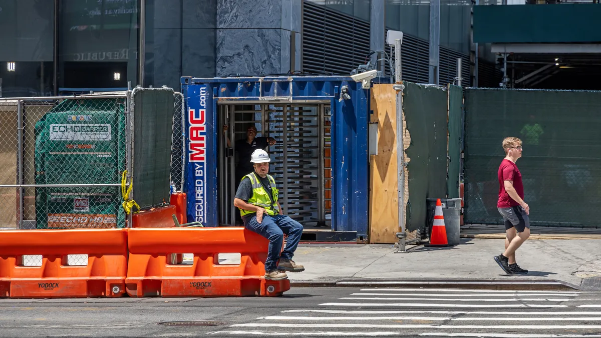 Construction worker wearing hardhat sits on barrier while a pedestrian walks by on a street.