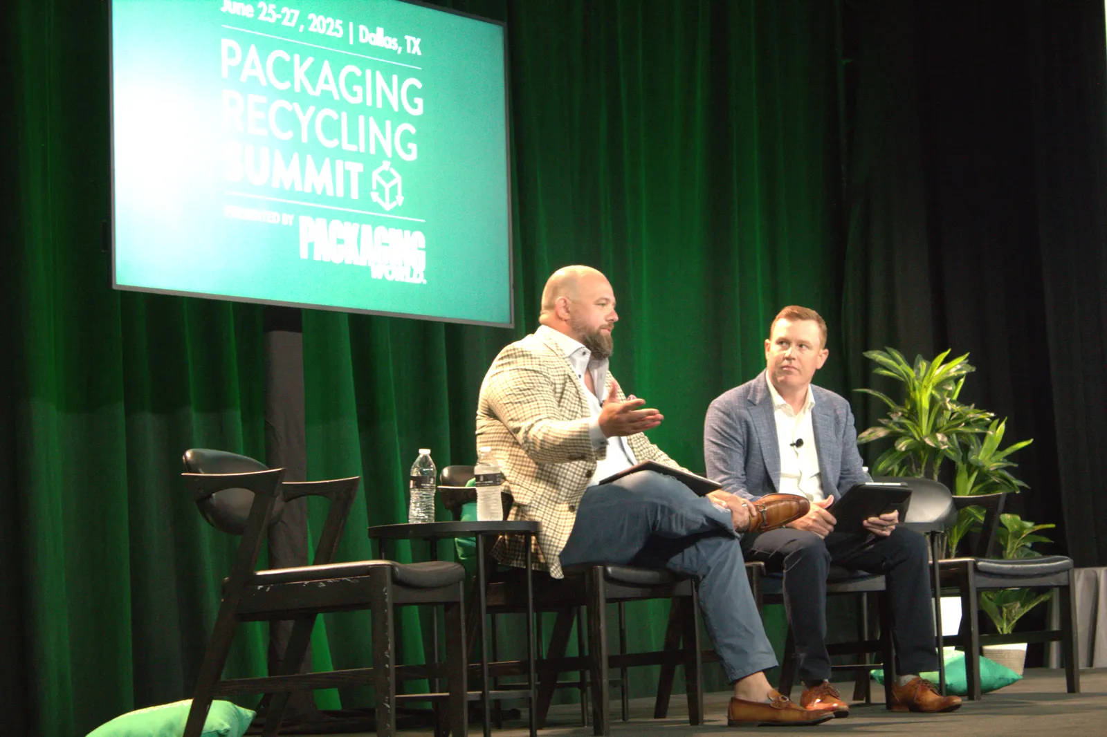 Two people sit in chairs on a stage in front of a green background that says Packaging Recycling Summit.