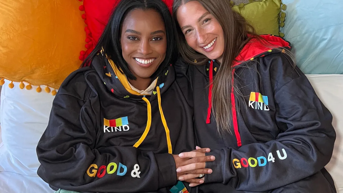 Ciara Miller and Amanda Batula lounge in bed wearing Kind hoodies.