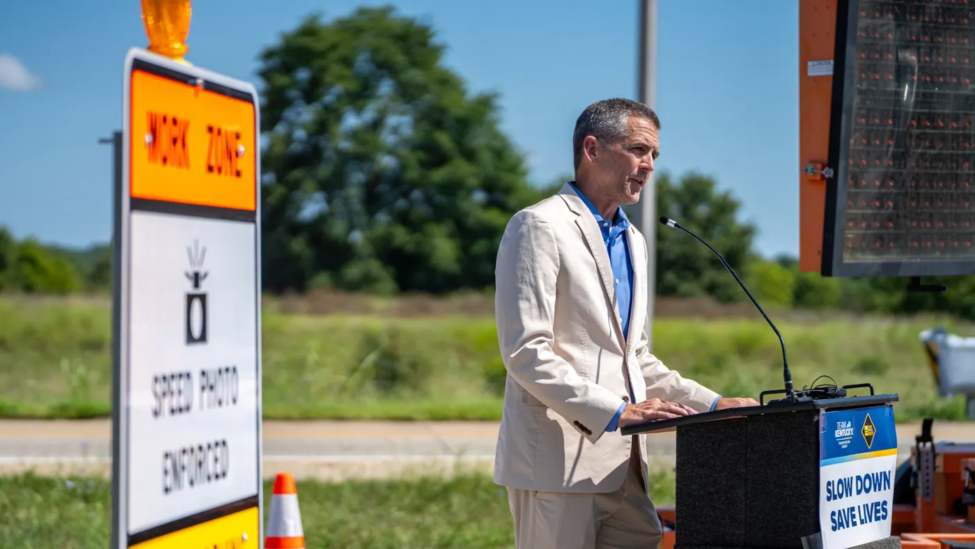 A man speaks at a podium next to road signs.