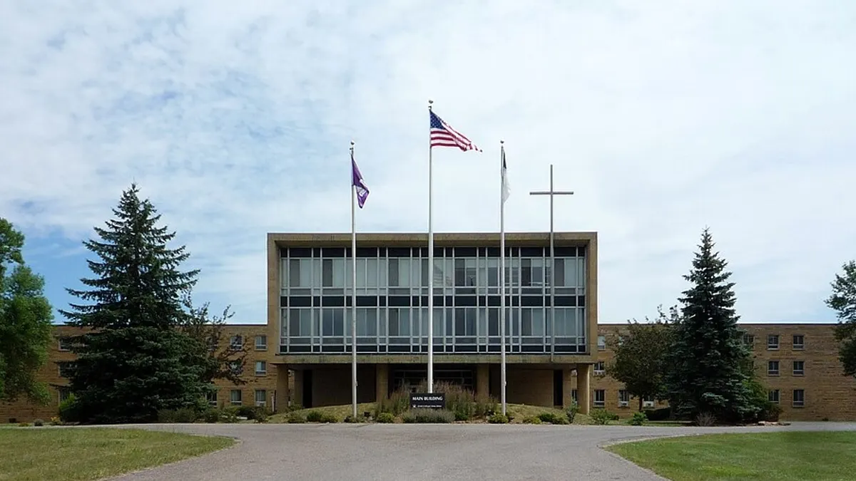 A campus building sits in the middle ground, behind a large cross and a flag pole with the U.S. flag.