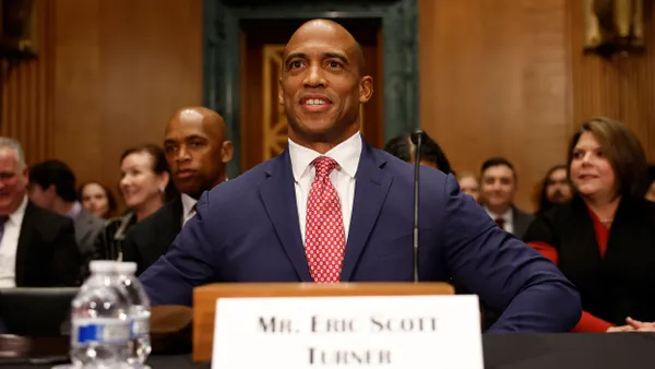 Seated man in dark blue suit with a microphone and a many other people seated behind him.