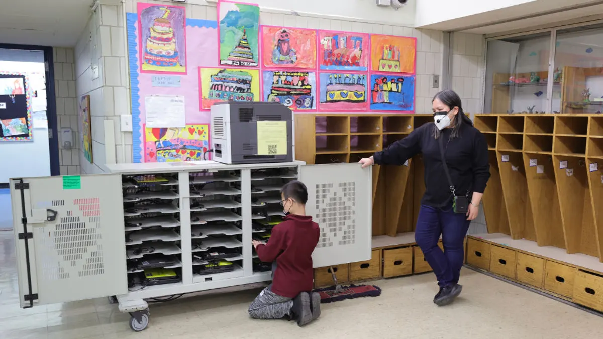 A student is near a cabinet holding laptops. A teacher stands nearby watching the student