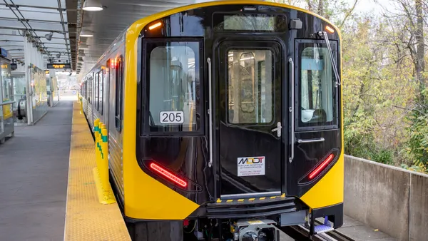 A head-on view of a stainless steel train painted yellow and black on the front end alongside a station platform.