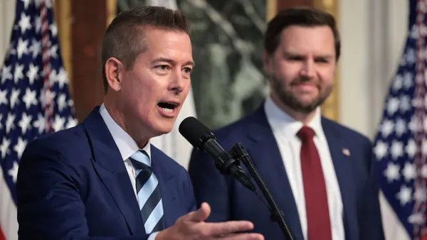 One man speaks at a microphone in front of flags and another man.