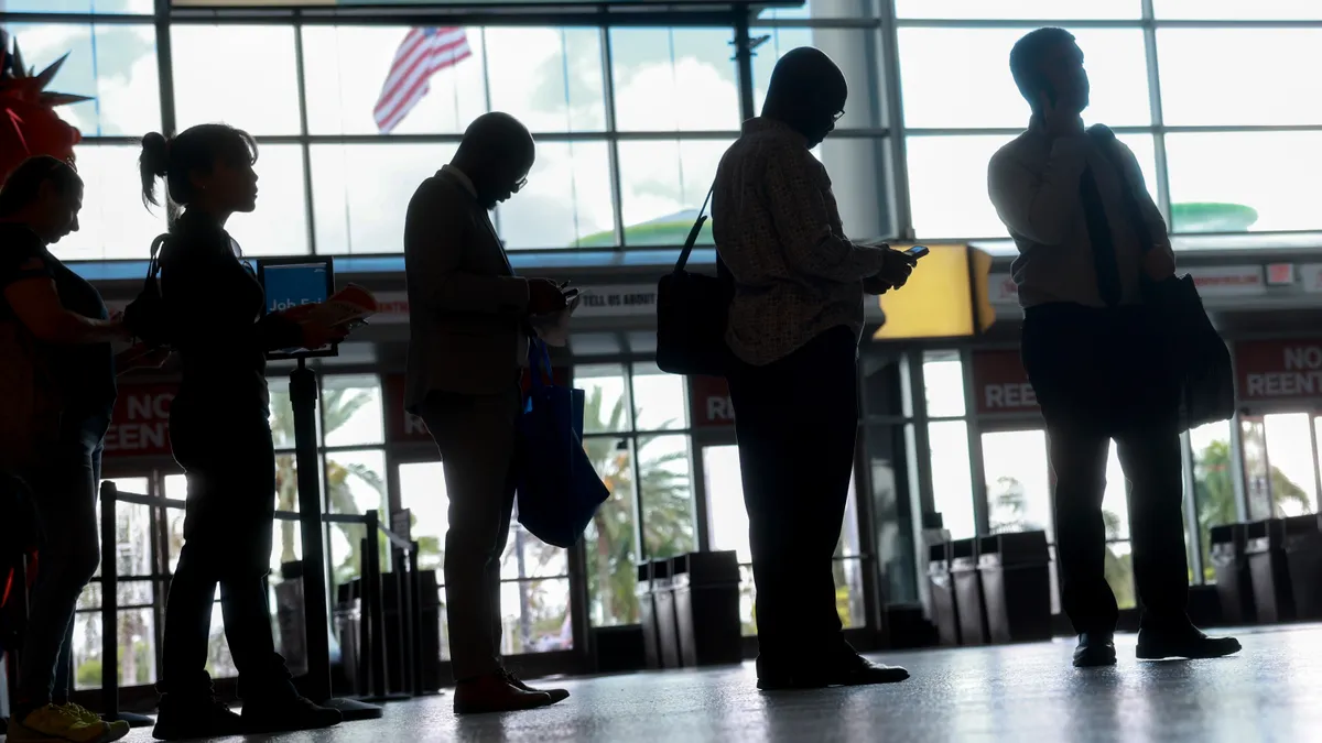 The silhouettes of individuals are seen standing in a line in front of a window at a job fair.