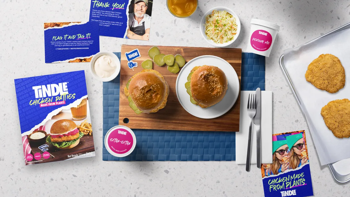 Components of Tindle's Motel Chicken Sandwich meal kit on a white table. Sandwiches are in the center on a cutting board, with packages, sauces, coleslaw, info cards and a baking tray with patties.