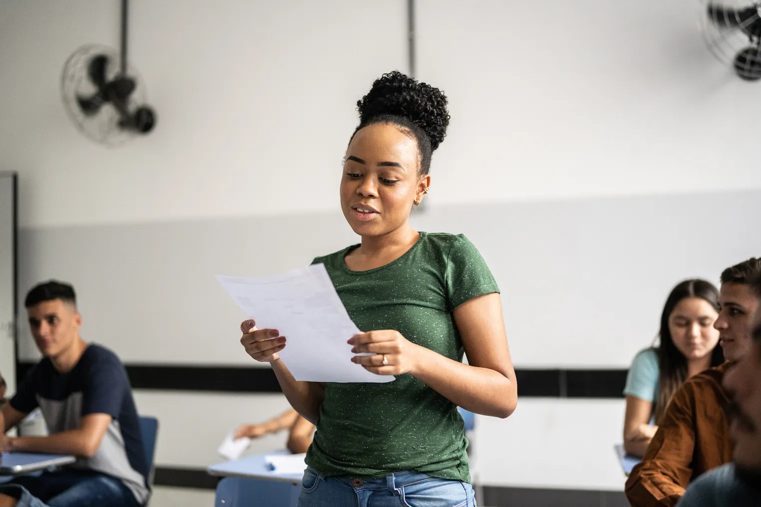 A Black teenage girl in a green shirt stands among the desks of her peers in a classroom and reads aloud from a sheet of paper.