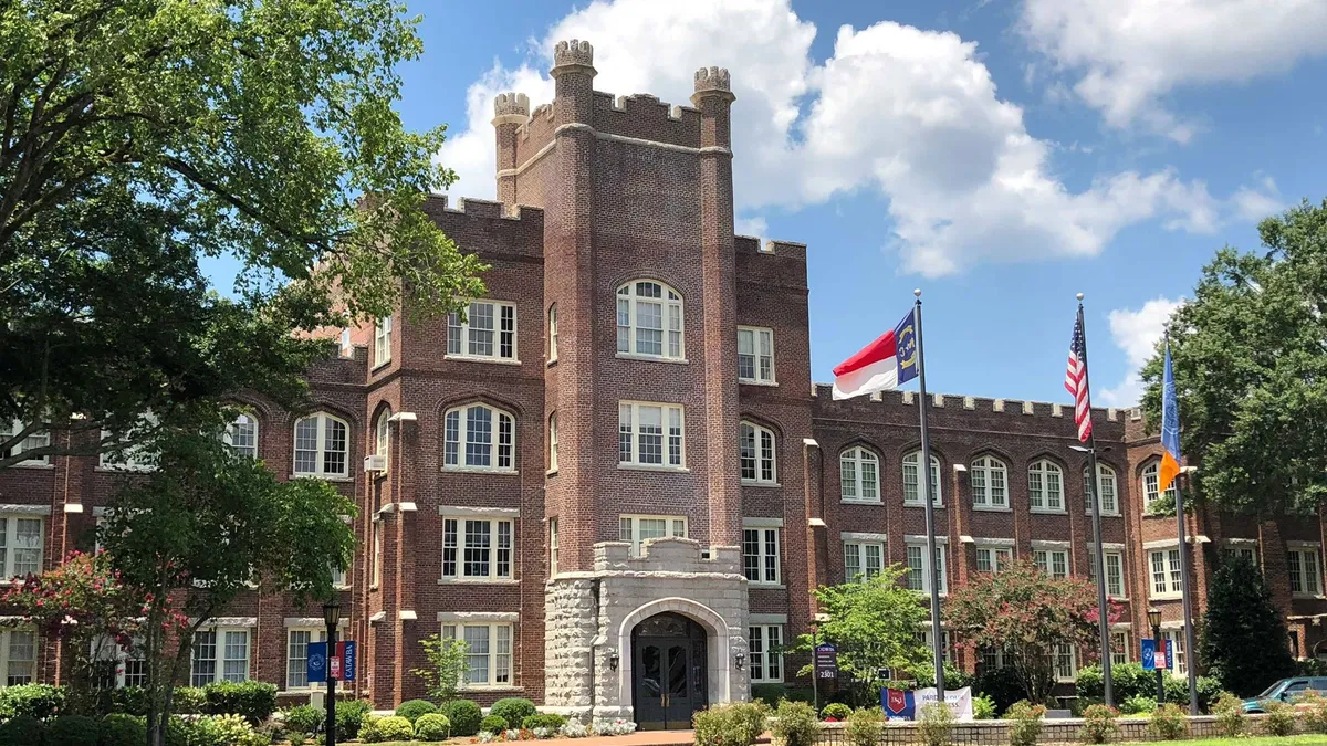 An academic building resembling a small Tudor castle stands behind a green grass lawn on a clear day.